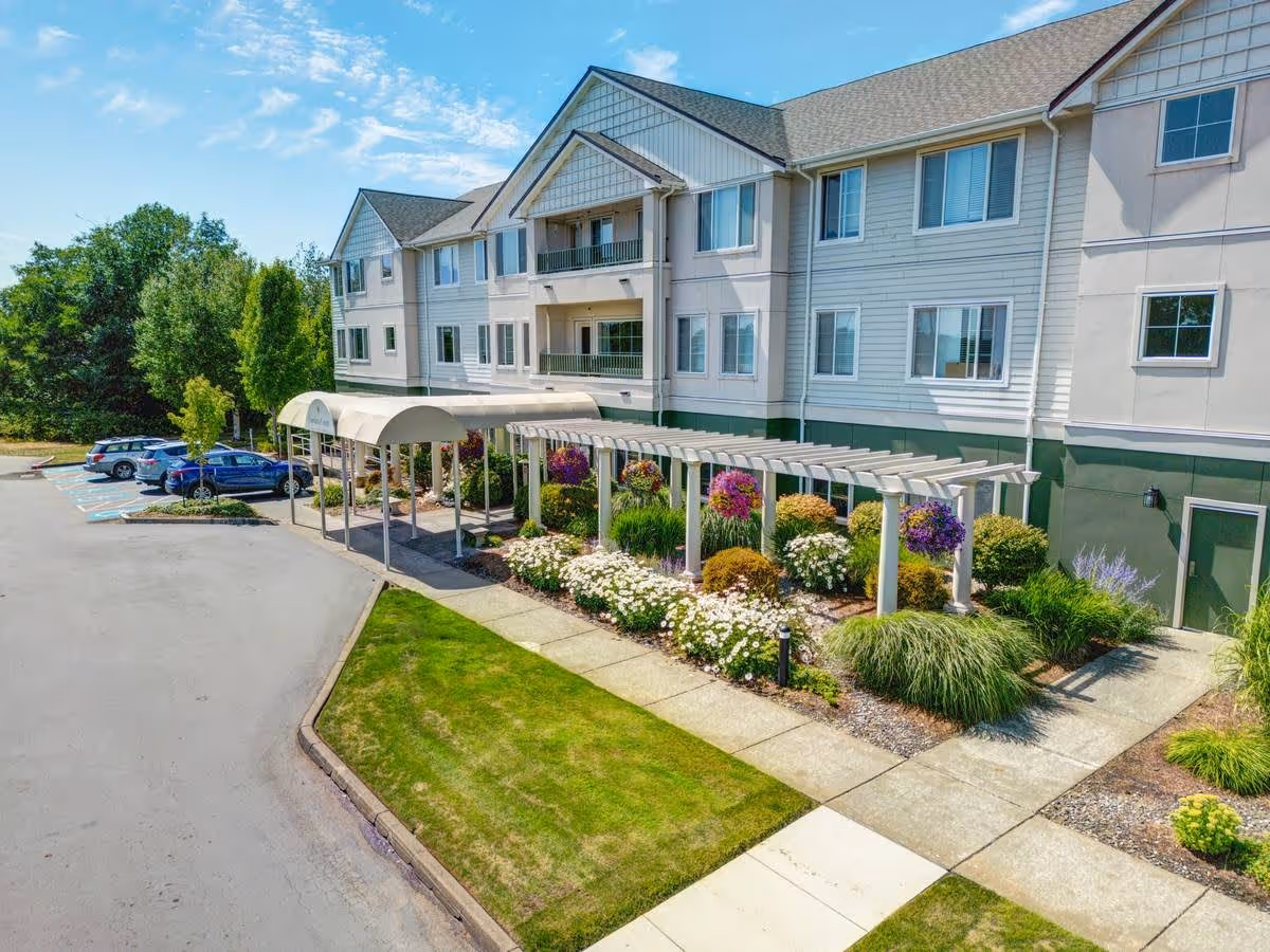 Exterior view of a multi-story senior living facility named Cordata Court with a covered entrance, landscaped garden with flowers and shrubs, a pergola with hanging flower baskets, and a parking area with several cars under a partly cloudy sky.