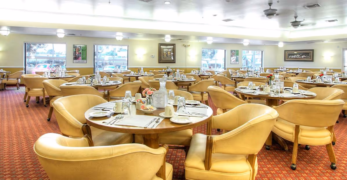 A spacious dining room with multiple round wooden tables set for meals, each surrounded by comfortable beige upholstered chairs. The tables have place settings including plates, cups, utensils, and small flower arrangements. Large windows allow natural light to fill the room, and the walls are decorated with framed artwork. The carpet is patterned in red tones, and ceiling fans are visible above.