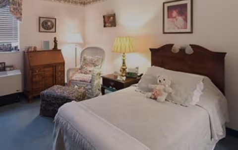 Cozy bedroom with a neatly made bed topped by a stuffed bear, a bedside table and lamp, a patterned armchair with ottoman, and a wooden dresser with framed pictures.