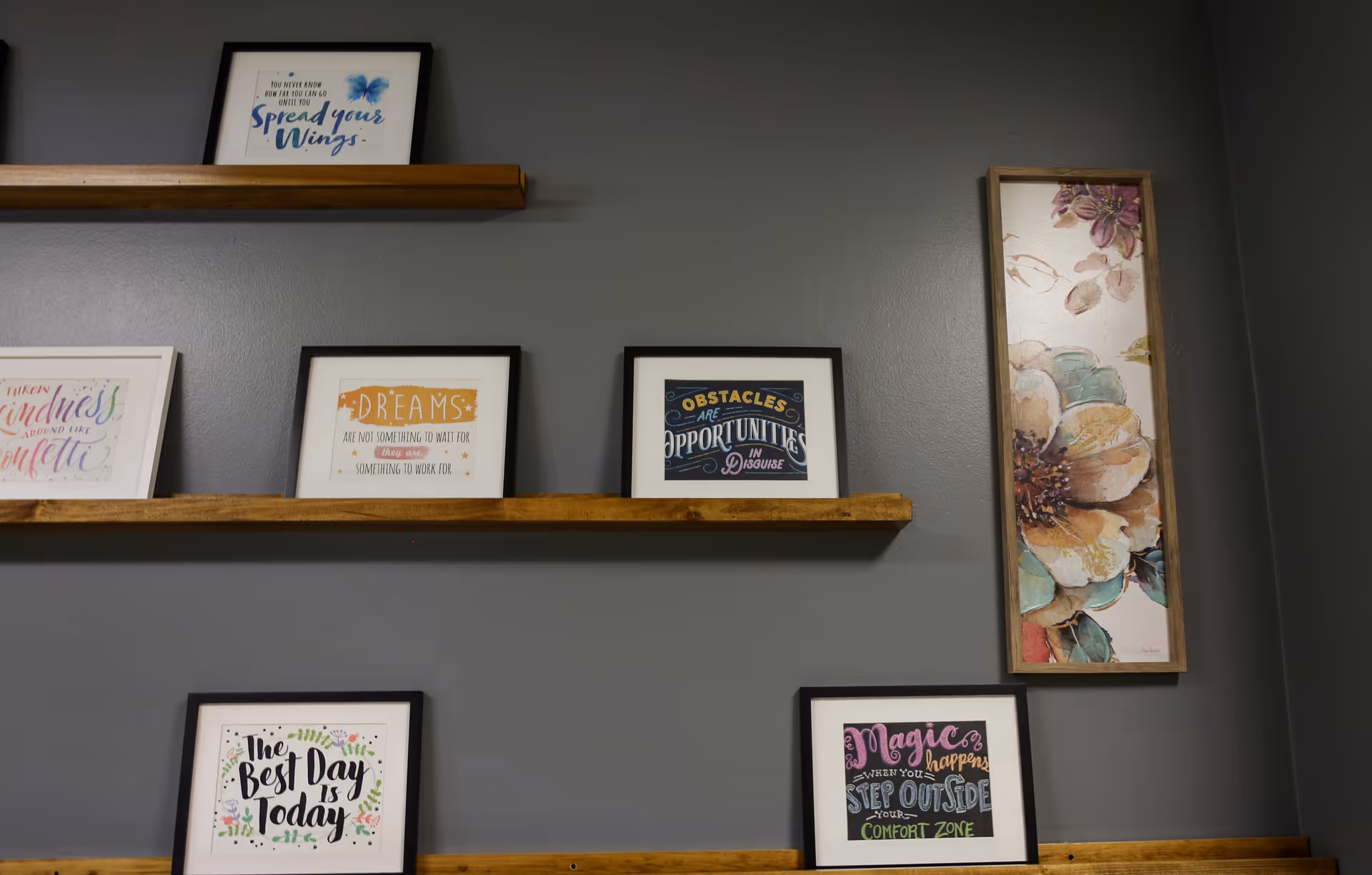 A gray wall with three wooden shelves displaying framed inspirational quotes and a floral painting. The quotes include messages like 'Spread your Wings,' 'Dreams are not something to wait for, something to work for,' 'Obstacles are opportunities in disguise,' 'The Best Day is Today,' and 'Magic happens when you step outside your comfort zone.'