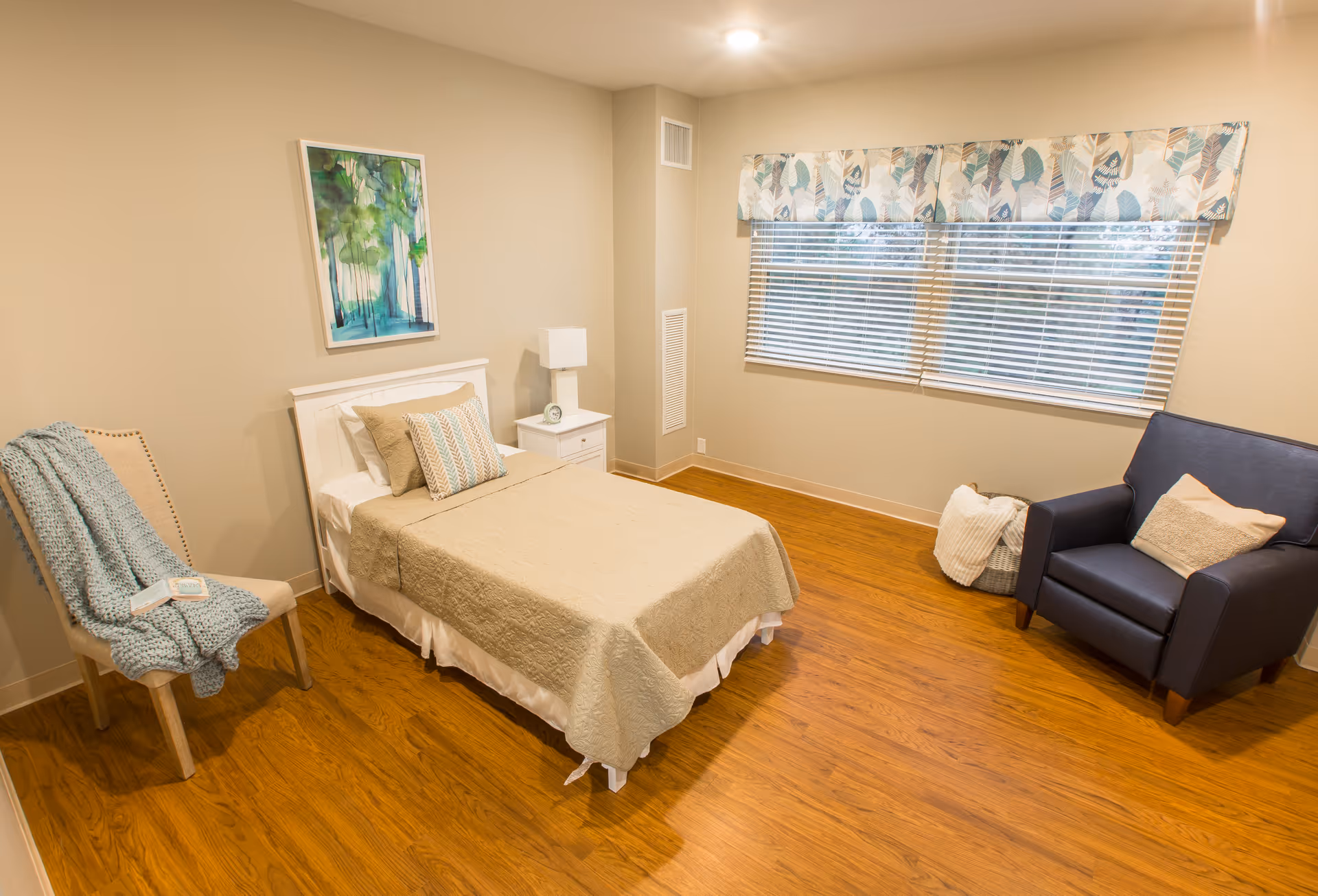 A cozy bedroom with a single bed covered in beige bedding and decorative pillows. There is a white nightstand with a lamp and clock next to the bed. A beige chair with a blue knitted blanket and a book is placed on the left side. On the right side, there is a dark blue armchair with a beige pillow and a basket next to it. The room has wooden flooring, beige walls, a large window with blinds and patterned valance, and a framed painting of trees on the wall above the bed.