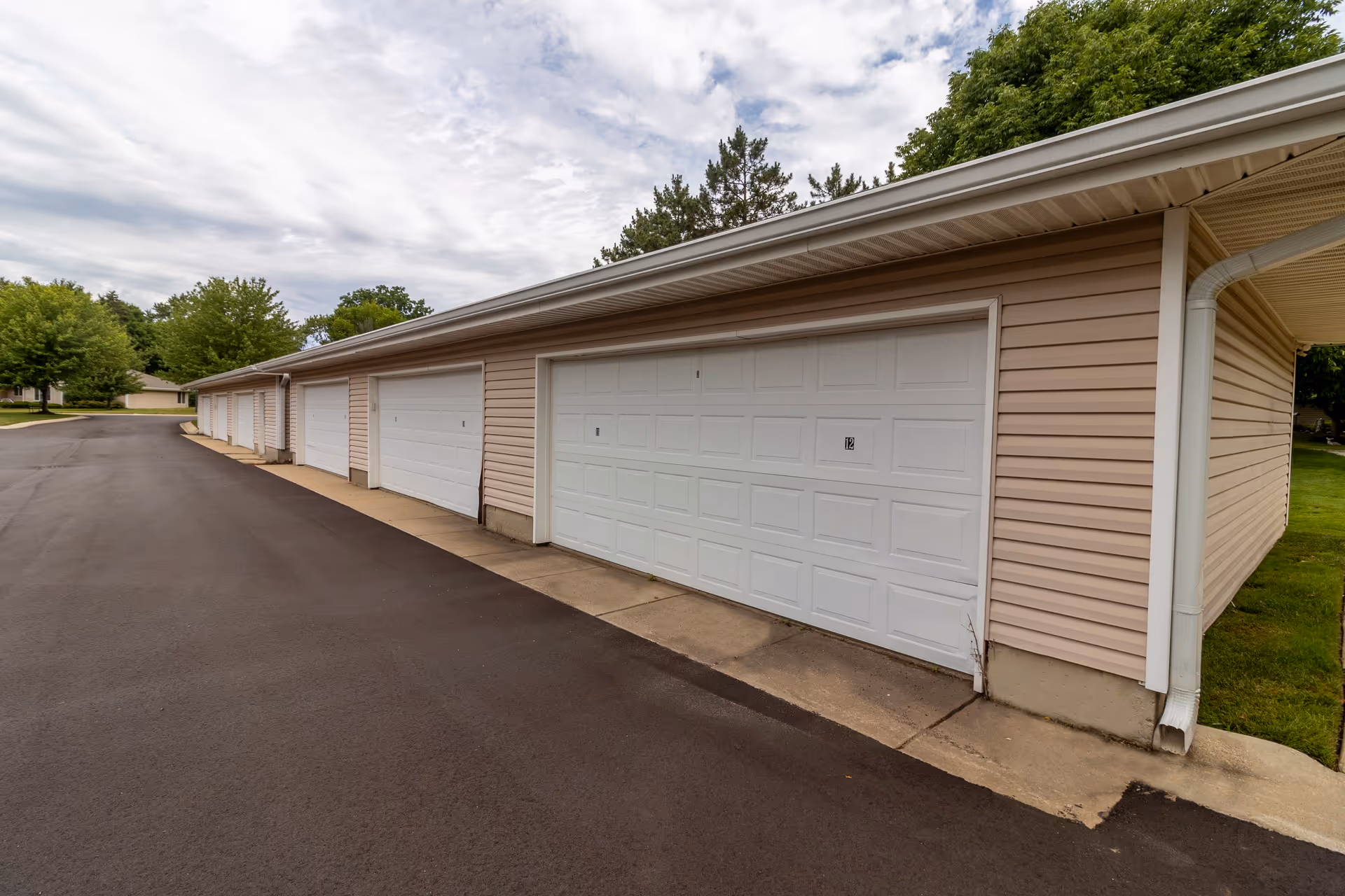 A row of beige garages with white garage doors along a paved driveway, surrounded by green trees and grass under a cloudy sky.