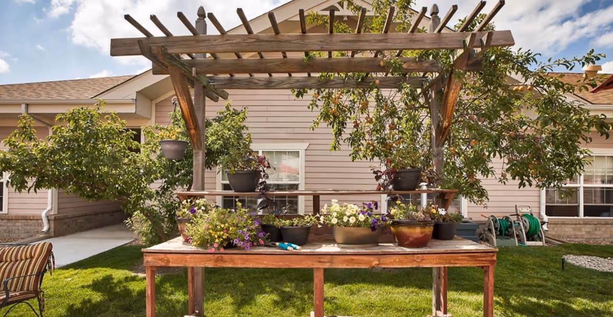 Outdoor garden area at Juniper Village at Aurora featuring a wooden pergola with various potted plants and flowers on a wooden table, surrounded by green grass and trees, with the facility building visible in the background under a partly cloudy sky.