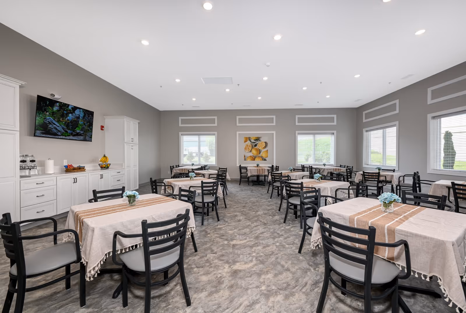 Bright communal dining room with multiple tables covered in beige tablecloths, black chairs, windows along the walls and a TV above a white service cabinet.