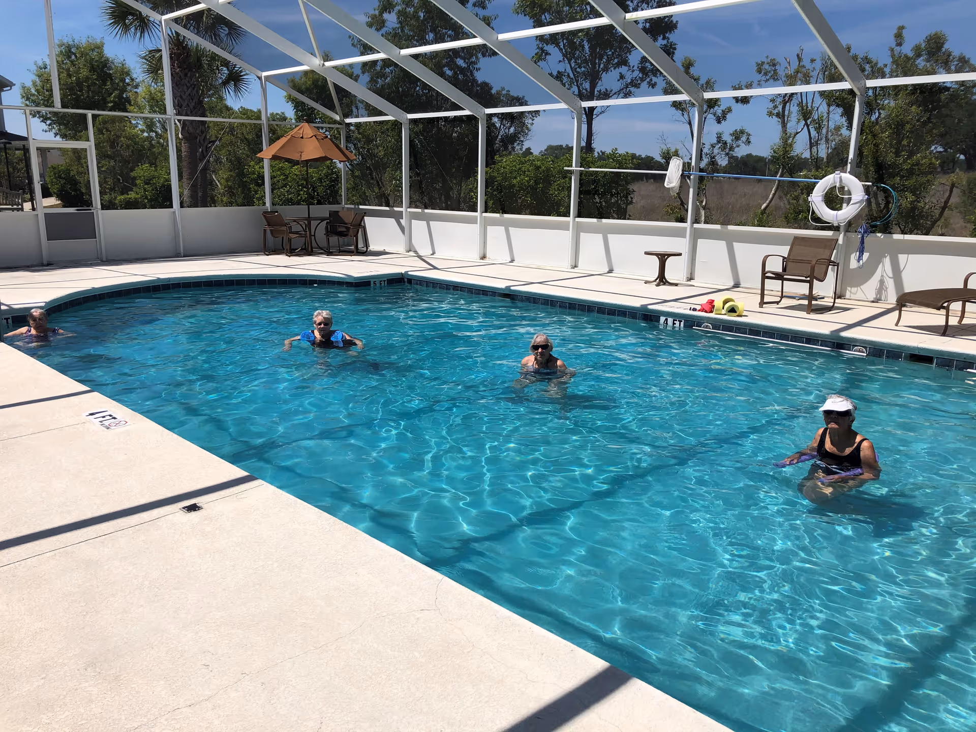 Four elderly women are enjoying a swim in a rectangular outdoor pool enclosed by a white screened structure. The pool area has a concrete deck with chairs, a table with an umbrella, and pool safety equipment such as life rings. Trees and greenery are visible outside the enclosure under a clear blue sky.