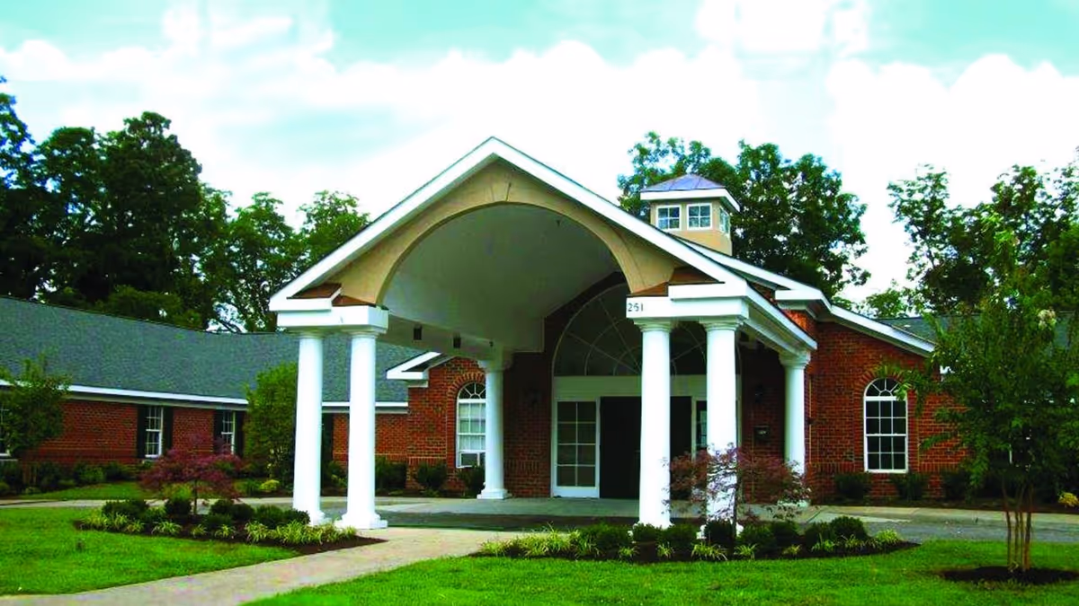 Front exterior view of a single-story brick building with white columns supporting a covered entrance. The building is surrounded by green grass, small trees, and shrubs under a partly cloudy sky.