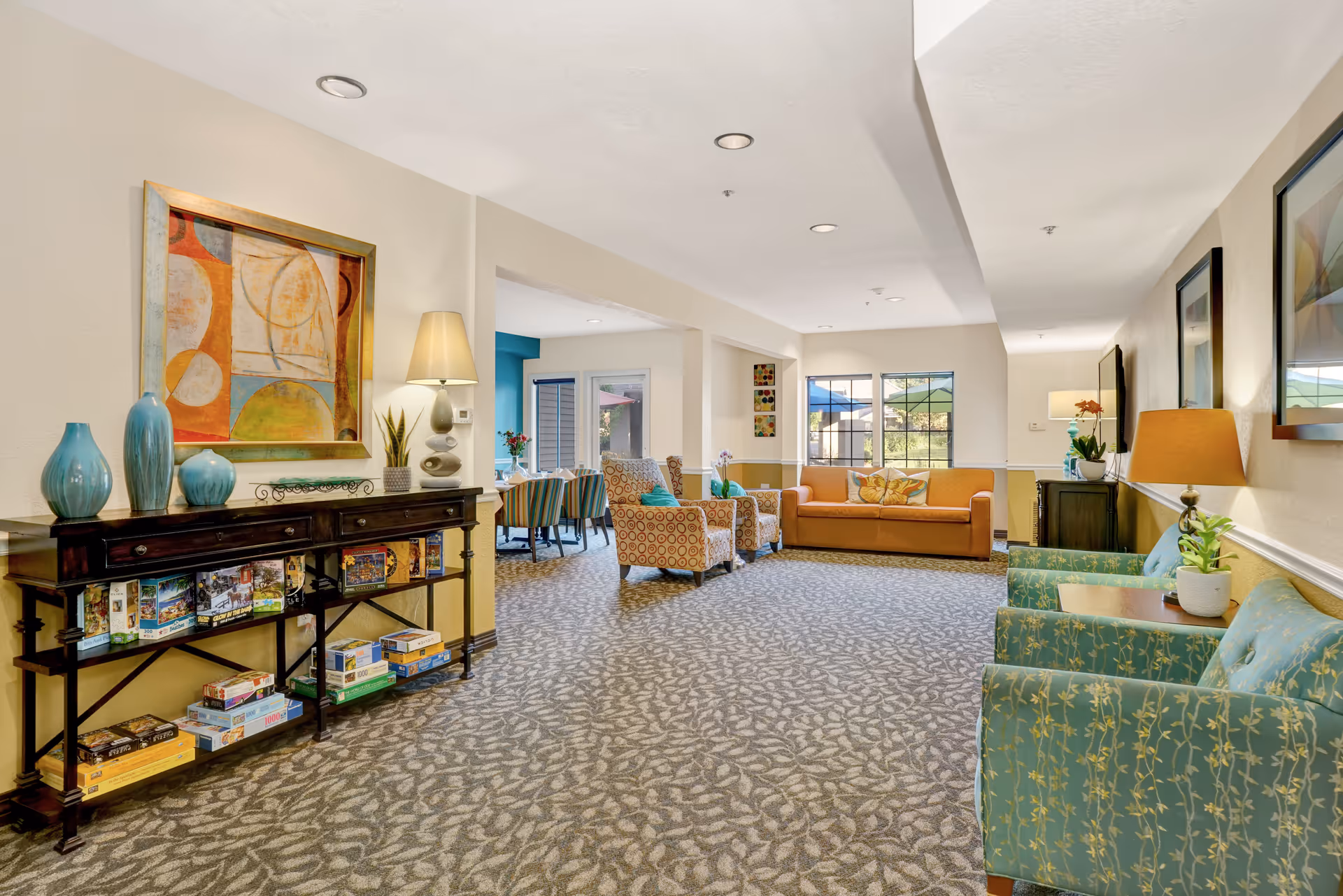 A bright and spacious senior living facility common area with patterned carpet flooring, multiple seating options including teal armchairs, patterned armchairs, and an orange sofa. The room features decorative vases on a dark wooden console table, framed abstract artwork on the walls, table lamps, and large windows letting in natural light. A dining area with a table and chairs is visible in the background.