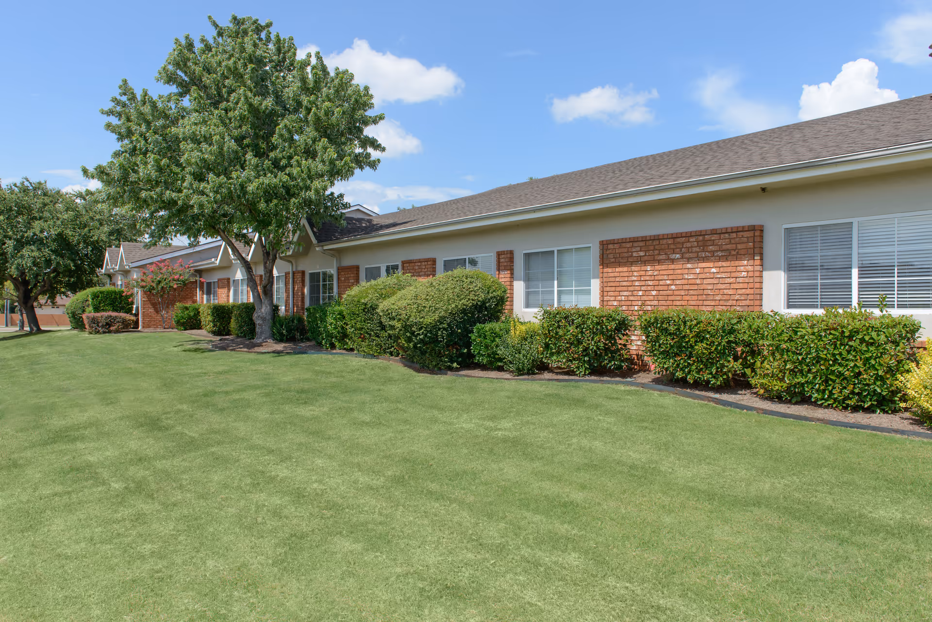 Single-story brick senior living building with a manicured lawn, shrubs, and trees under a blue sky.