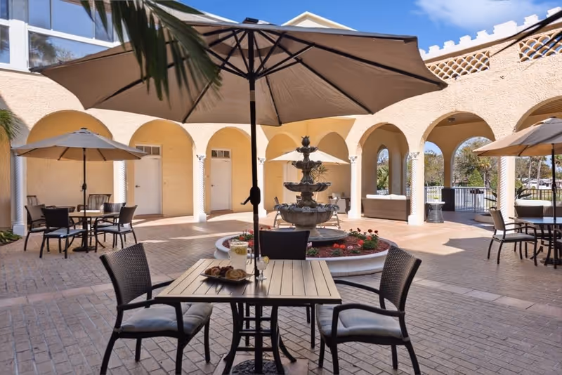 Outdoor courtyard area at Riviera Senior Living with several tables and chairs under large umbrellas, a central decorative fountain surrounded by flowers, and arched walkways in the background.
