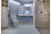 A spacious, accessible bathroom with a white toilet, a wall-mounted sink with exposed plumbing, a large mirror above the sink, and a walk-in shower with a handheld showerhead and grab bars. The walls are light blue and the shower area has beige tiles.