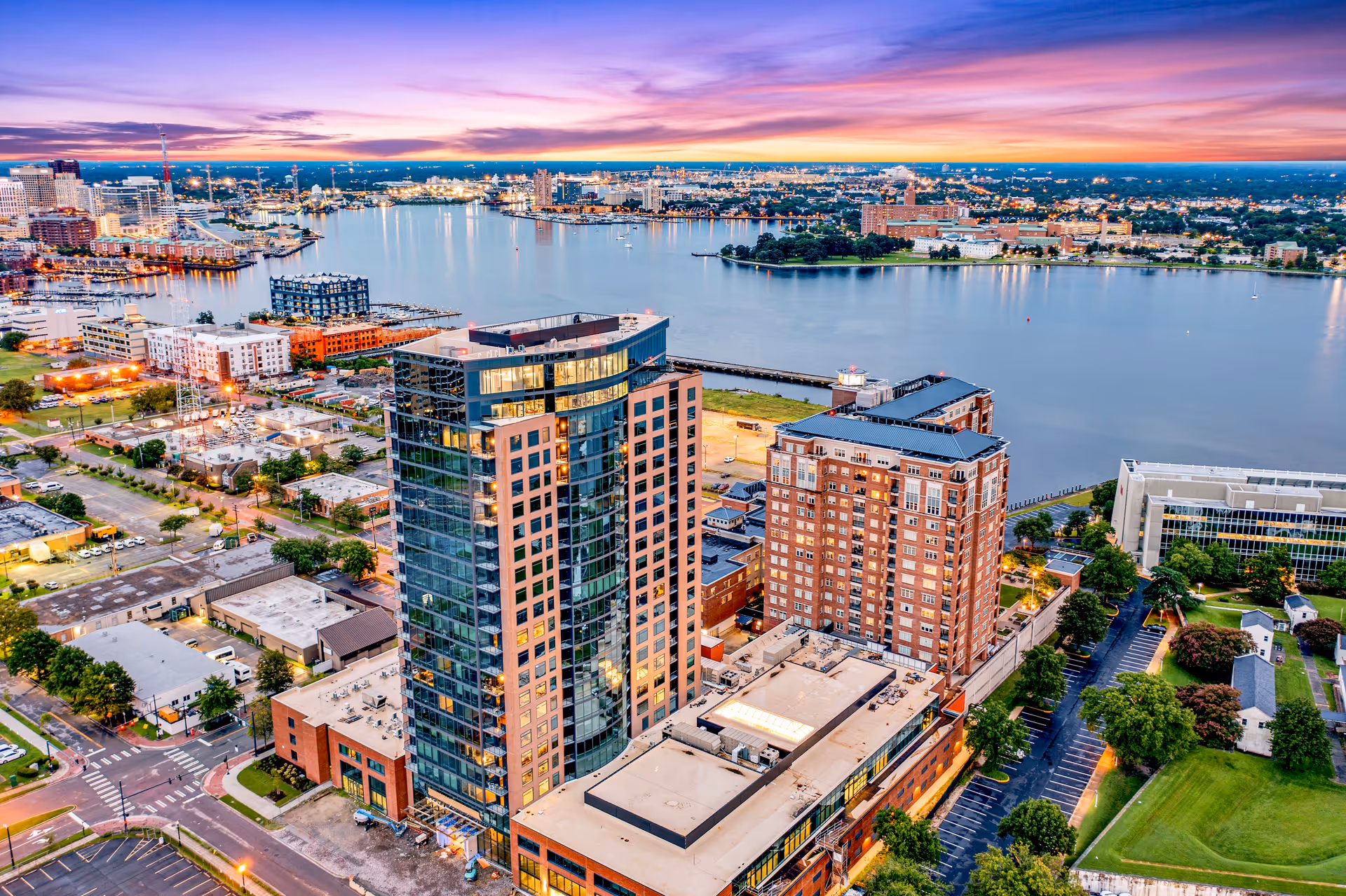 Aerial view of a waterfront cityscape at sunset featuring tall buildings, a river, and a vibrant sky with purple and orange hues.