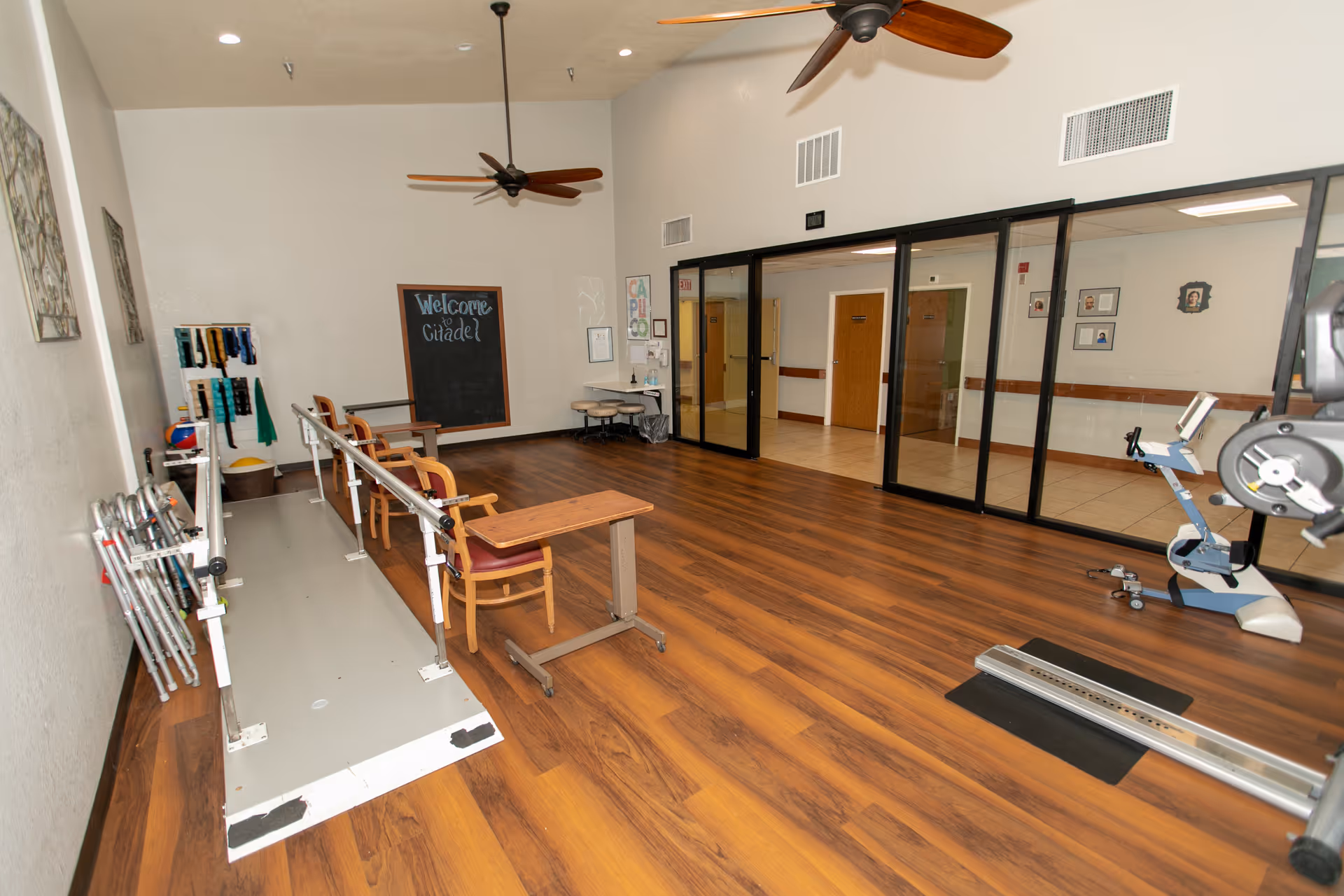 A rehabilitation or physical therapy room with wooden flooring, parallel bars for walking exercises, chairs with attached tables, exercise equipment, and a chalkboard on the wall that says 'Welcome Citadel'. The room has ceiling fans and large glass windows looking into an adjacent hallway.