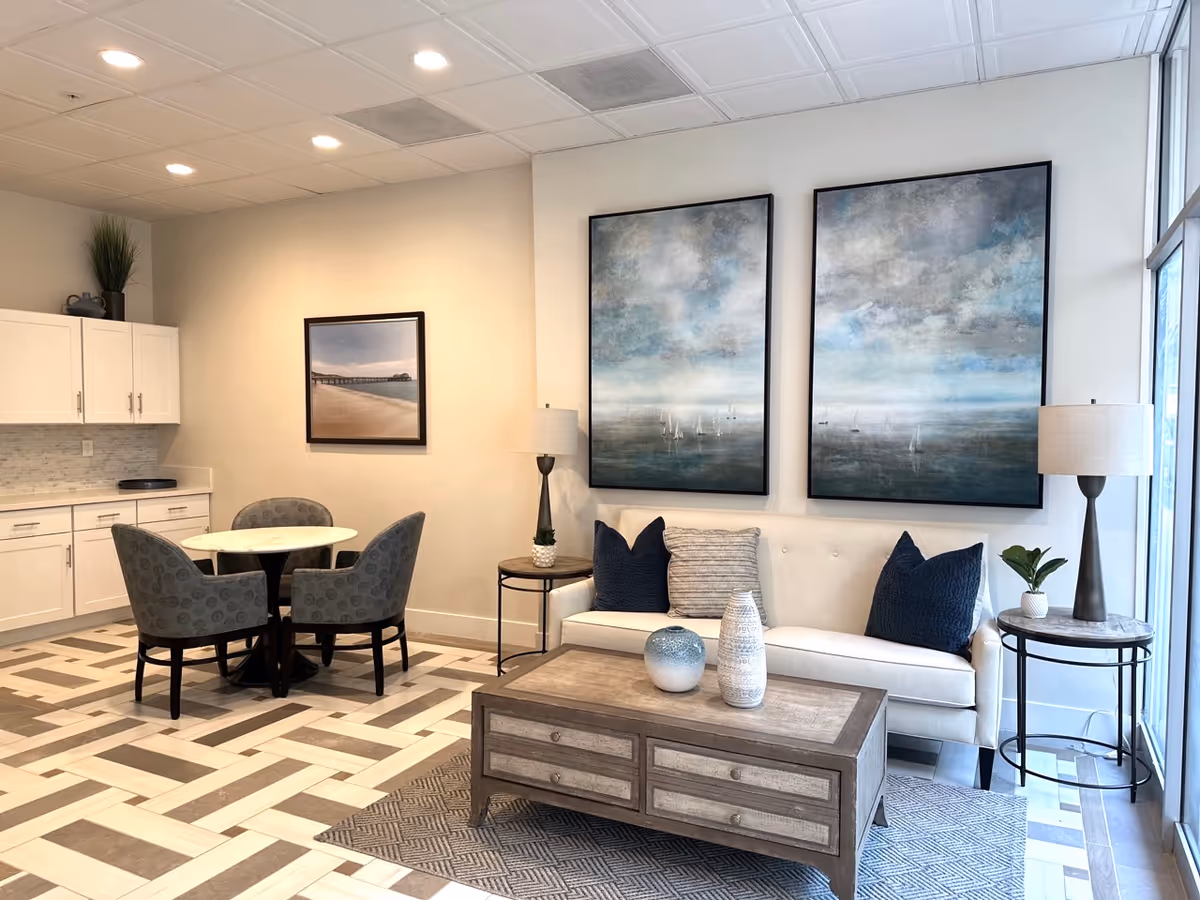 A cozy interior common area featuring a white sofa with three pillows, two large abstract paintings of a cloudy sky and sea above the sofa, a wooden coffee table with decorative vases, two side tables each with a lamp and a small plant, a round dining table with four upholstered chairs, white cabinetry with a countertop, and a patterned tile floor.