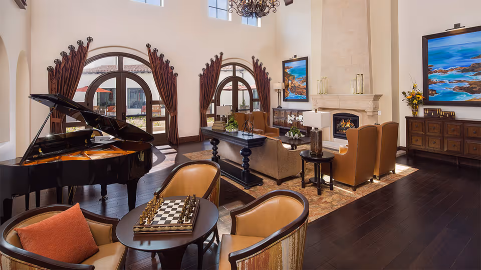 A spacious and elegant living room with high ceilings, large arched windows with brown curtains, a grand piano, a chess table with two chairs, a seating area with sofas and armchairs around a fireplace, and colorful ocean-themed paintings on the walls.
