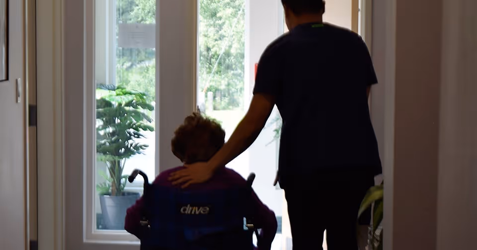 A caregiver gently guiding an elderly person in a wheelchair down a hallway towards a glass door with plants visible outside.