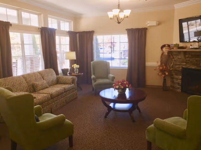 A cozy living room with a floral patterned sofa, three green armchairs, a round wooden coffee table with a flower arrangement, a stone fireplace with decorations, large windows with brown curtains, and a ceiling light fixture.
