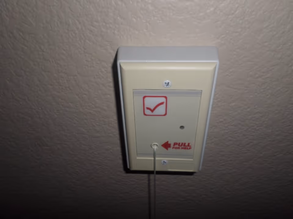 A beige emergency pull cord alarm mounted on a textured wall, with a red checkmark symbol and the text 'PULL FOR HELP' next to the cord.