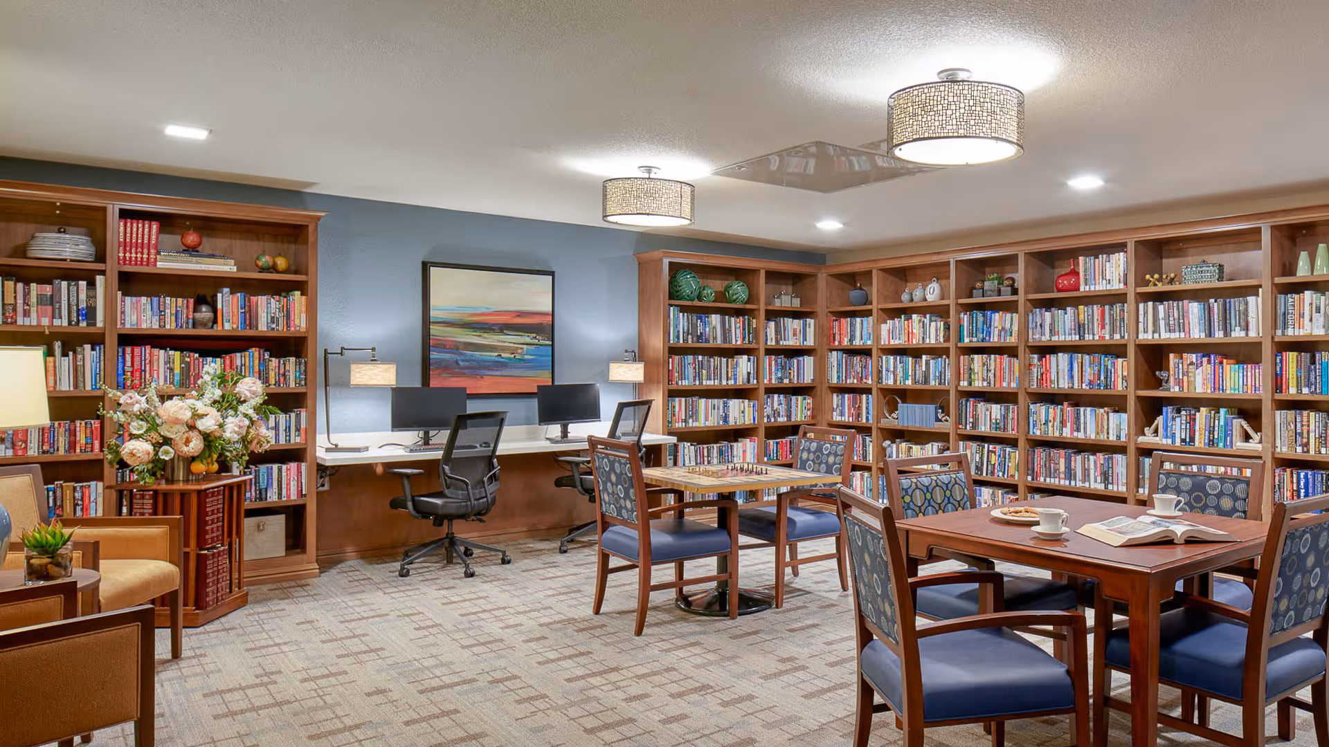 A cozy library room with wooden bookshelves filled with books lining the walls. There are two computer workstations with black office chairs against a blue accent wall. In the center, there are wooden tables with blue cushioned chairs, one table has an open book and a cup on it. The room is well-lit with ceiling lights and decorated with a floral arrangement and framed artwork.
