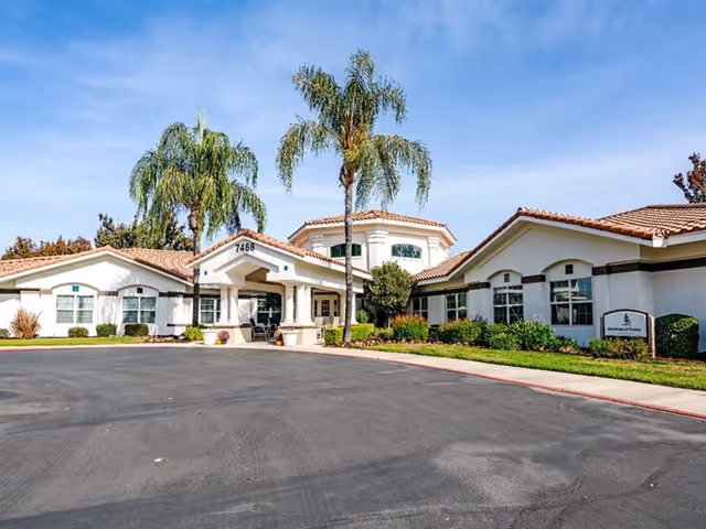 Exterior view of a single-story senior living facility with white walls and a tiled roof, surrounded by palm trees and landscaping under a clear blue sky. The entrance has a covered drop-off area with the number 7468 displayed above it.