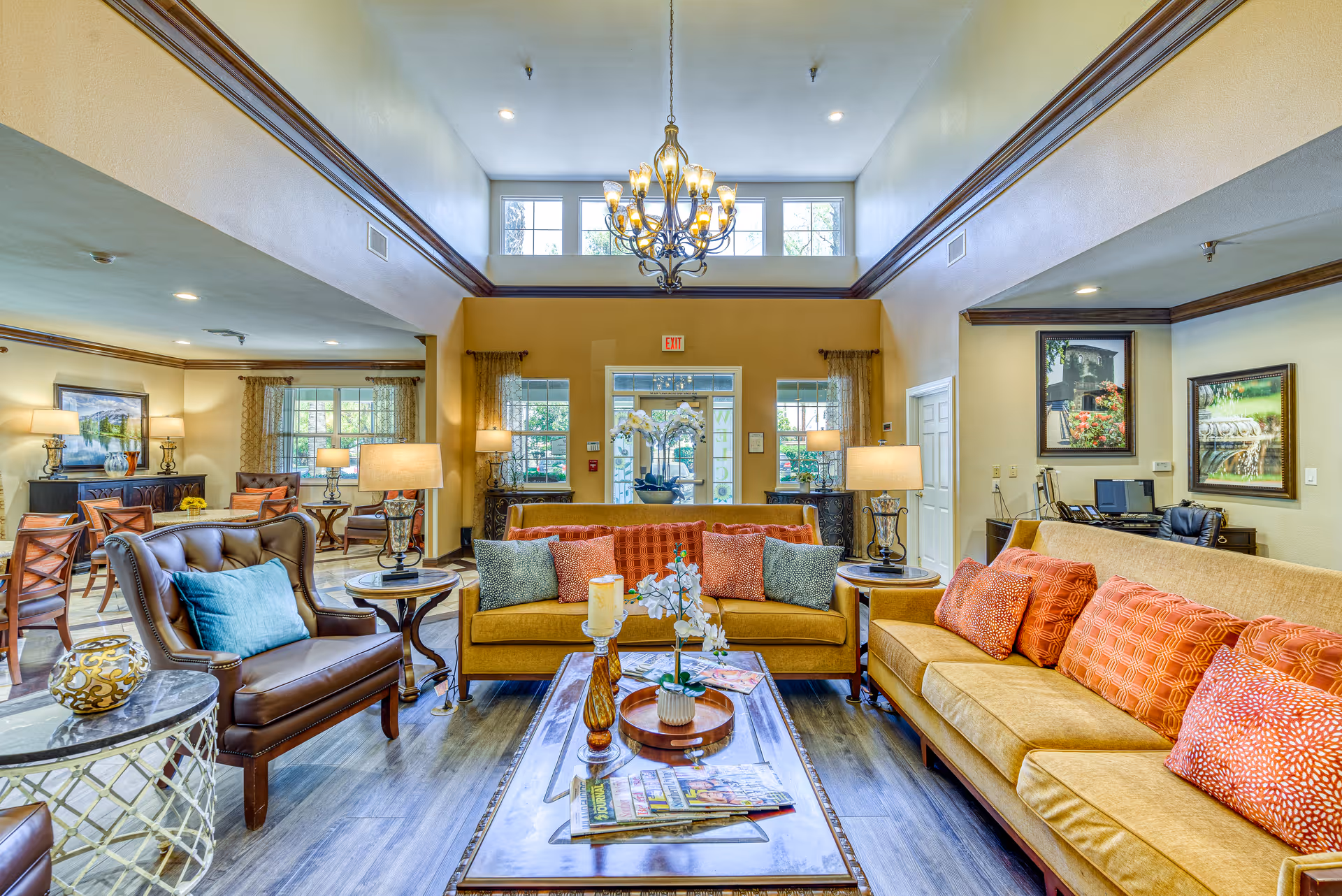 A spacious and well-lit living room area in a senior living facility with two mustard yellow sofas adorned with orange and patterned cushions, a brown leather armchair with a blue cushion, and a wooden coffee table with decorative items and magazines. The room features large windows, a chandelier hanging from a high ceiling, multiple lamps, framed artwork on the walls, and a view of the entrance door with glass panels.