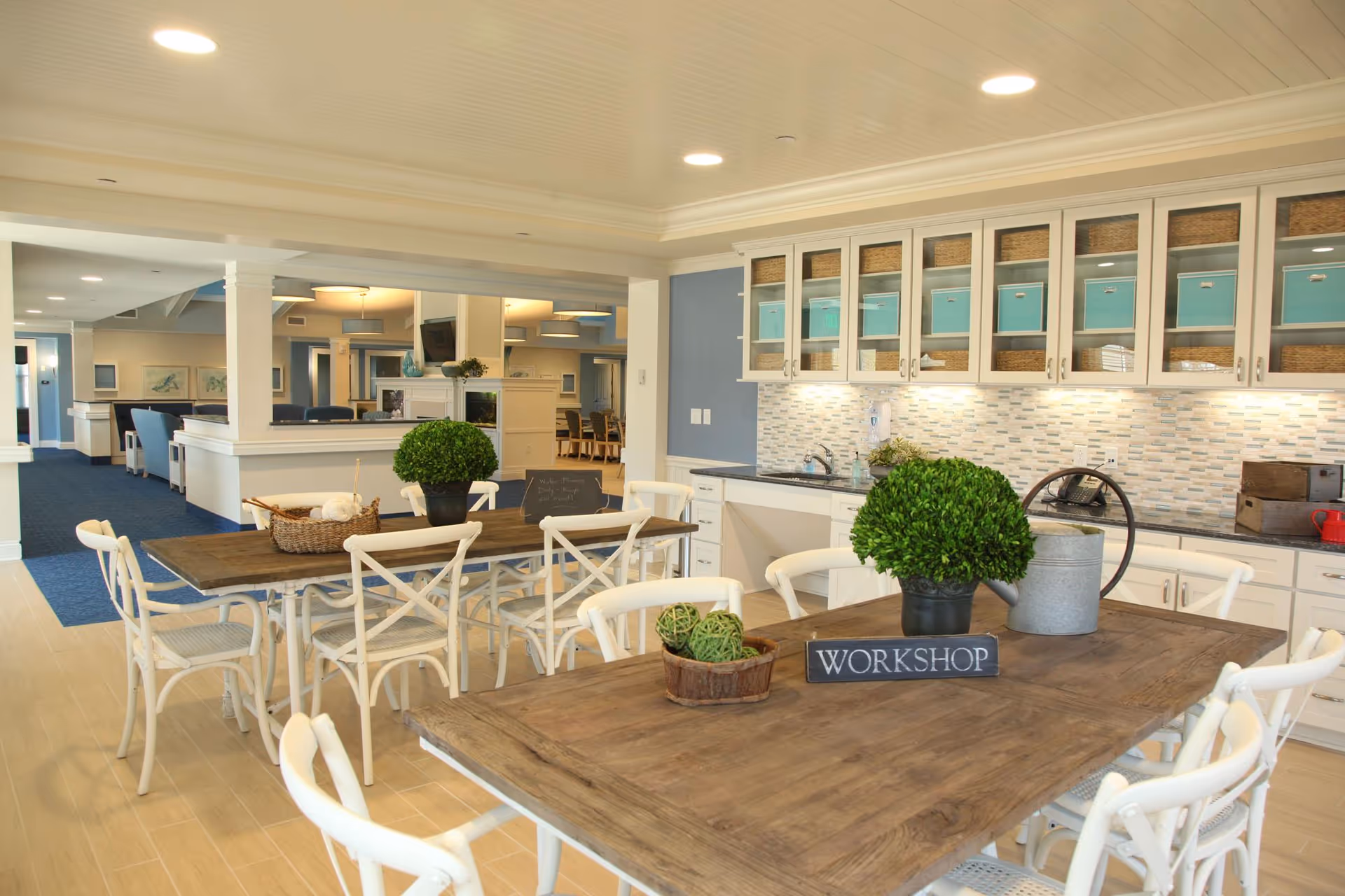Bright communal dining and kitchen area with wooden tables, white chairs, upper cabinets with turquoise bins, and a small sign reading 'WORKSHOP'.