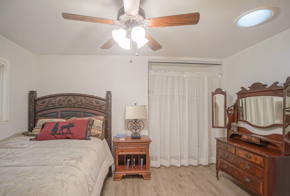 Well-lit bedroom with a carved wooden bed, matching dresser and nightstand, ceiling fan, and sheer curtains covering a closet or window.
