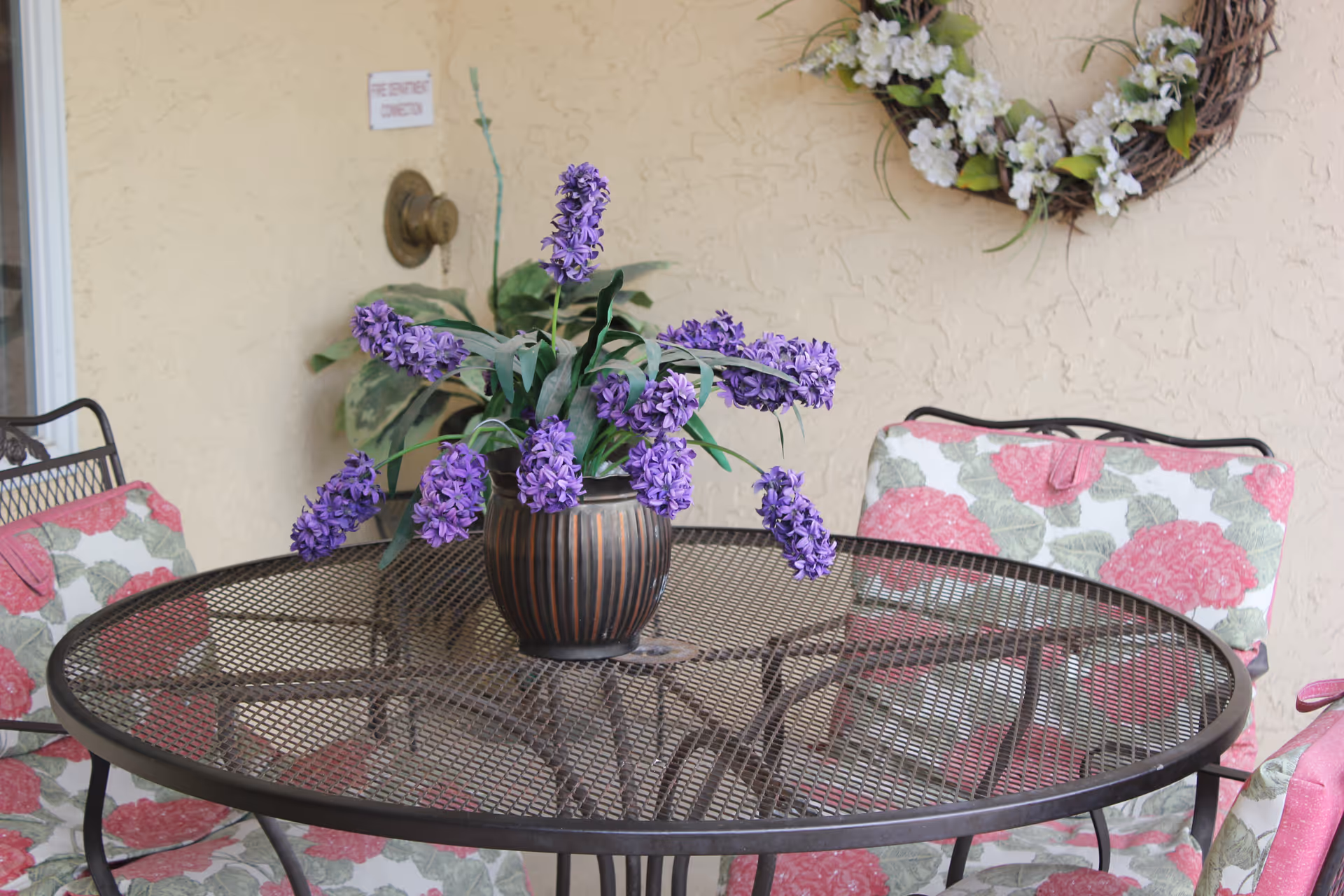 A round metal mesh outdoor table with a brown vase holding purple flowers in the center. Surrounding the table are chairs with floral cushions featuring pink and green patterns. A decorative wreath with white flowers hangs on a beige textured wall in the background.
