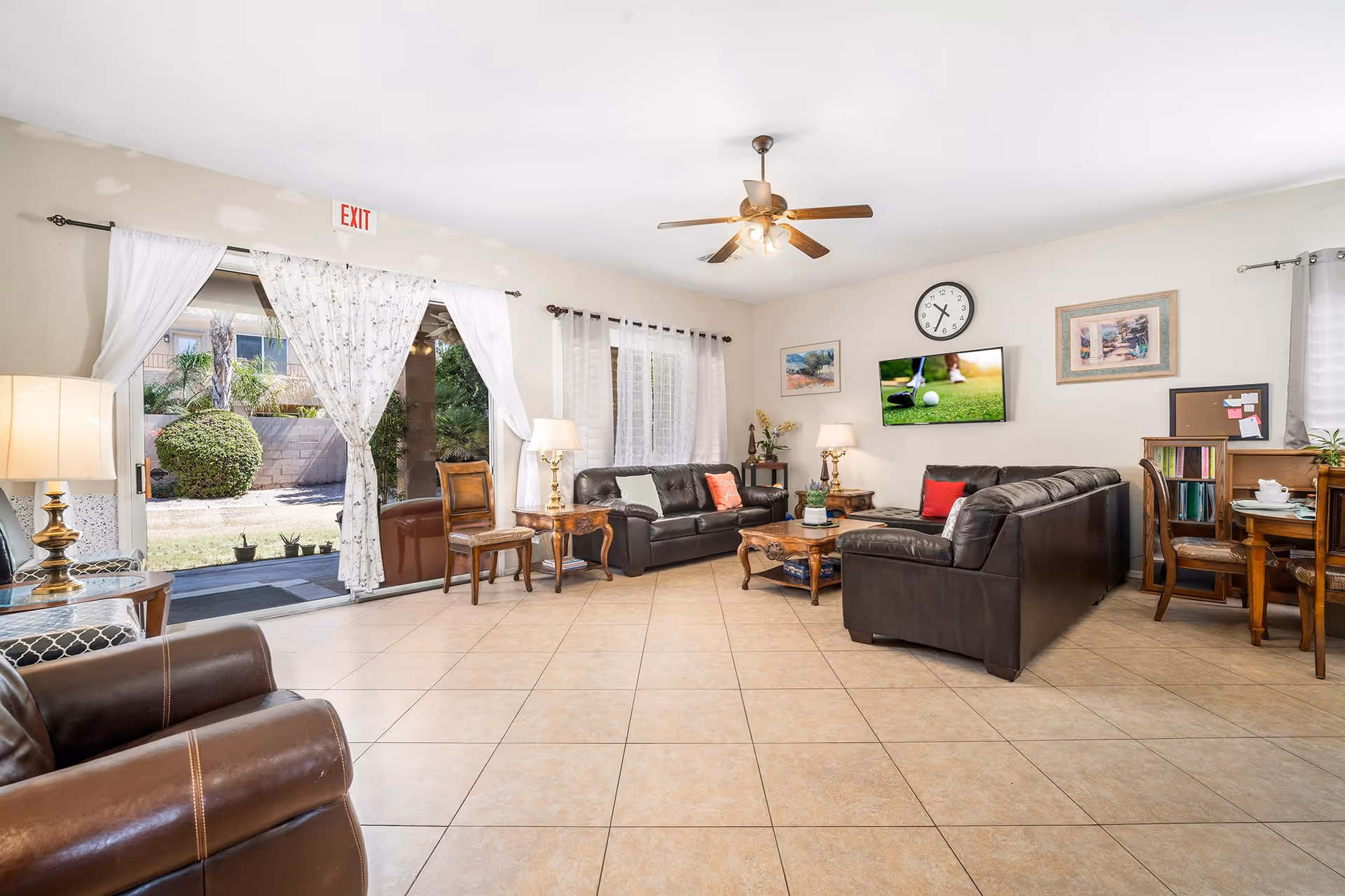 Bright communal living room with leather sofas and chairs, coffee tables, a wall-mounted TV and clock, and sliding glass doors opening to an outdoor area.