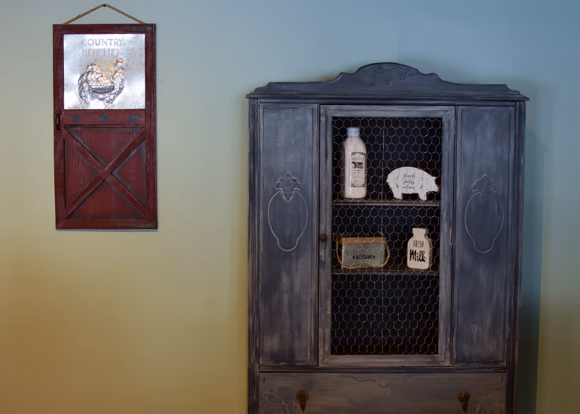 A rustic kitchen-themed wall decoration with a red wooden sign featuring a metal rooster and the words 'Country Kitchen' hanging on a light green wall next to a vintage gray cabinet with a wire mesh door displaying decorative items including a bottle, a pig-shaped sign, a small box, and a 'Fresh Milk' sign.