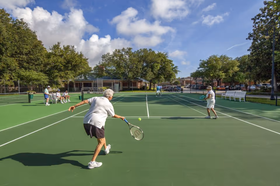 Two elderly women playing tennis on a green outdoor tennis court with a few other people sitting and standing nearby. Trees and buildings are visible in the background under a partly cloudy blue sky.