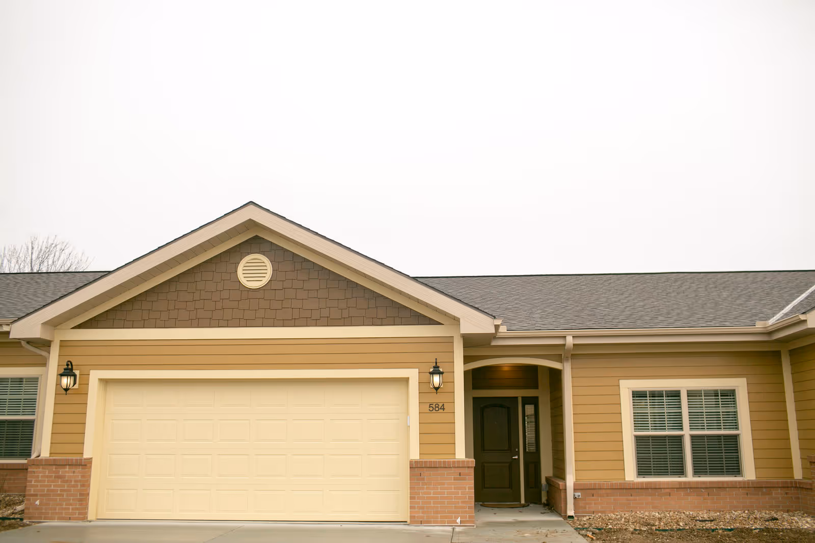 Exterior front view of a single-story residential building with a beige garage door, a brown front door under a small covered porch, two wall-mounted lantern lights, and windows with white blinds. The building has tan siding with brick accents at the base and a gray shingled roof.