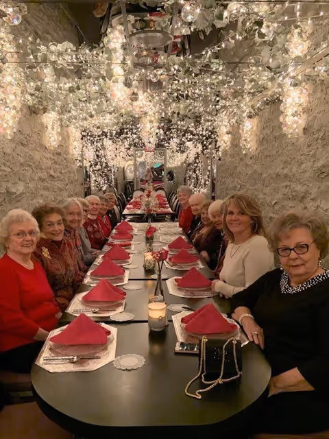 A group of elderly women seated around a long dining table set with red napkins, plates, and candles in a warmly lit room decorated with hanging white flowers and string lights.