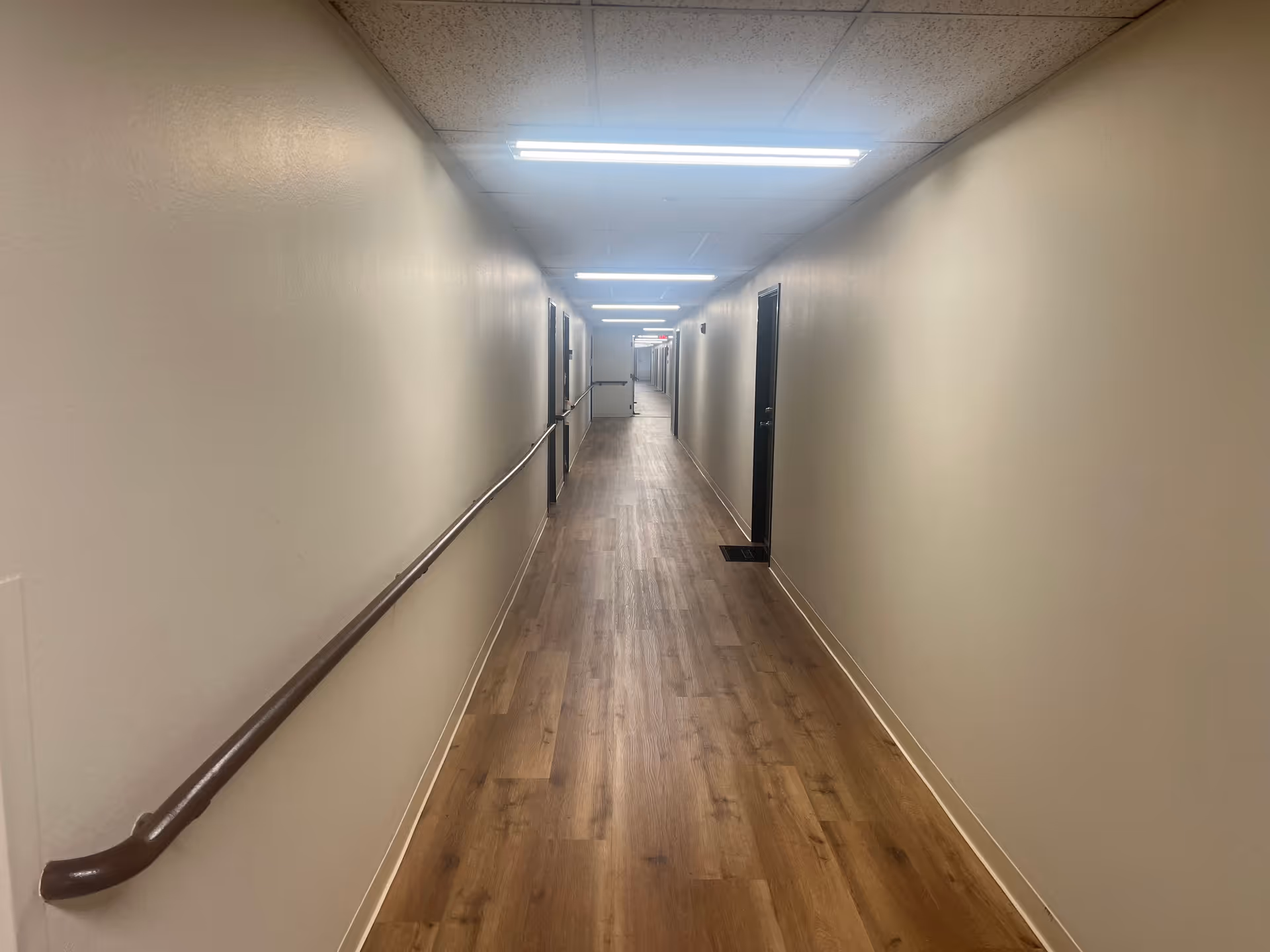Long narrow indoor corridor with wood-look flooring, handrails along the left wall, doors on the right, and fluorescent lights overhead.