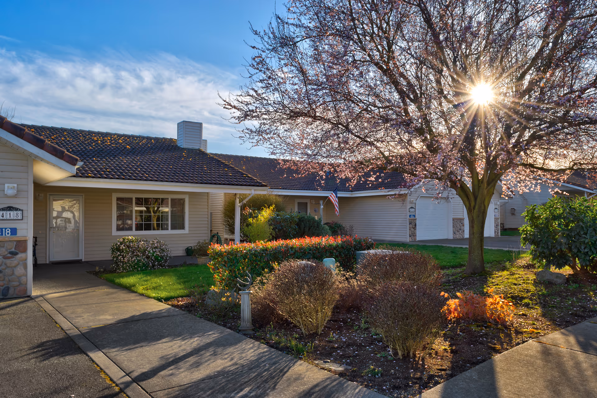 Exterior view of a single-story residential building with a tiled roof, a well-maintained garden with bushes and a tree with sunlight shining through its branches, and a concrete walkway leading to the entrance.