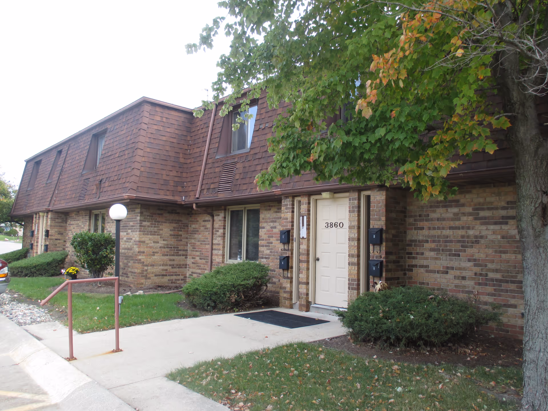 Front exterior of a two-story brick apartment building with a door numbered 3860, shrubs, sidewalk and a tree.