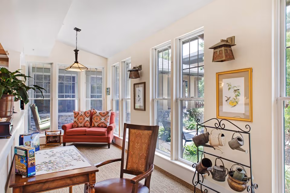 Bright sunroom-style sitting area with a red loveseat, wooden table and chairs, large windows, and a decorative rack of watering cans.