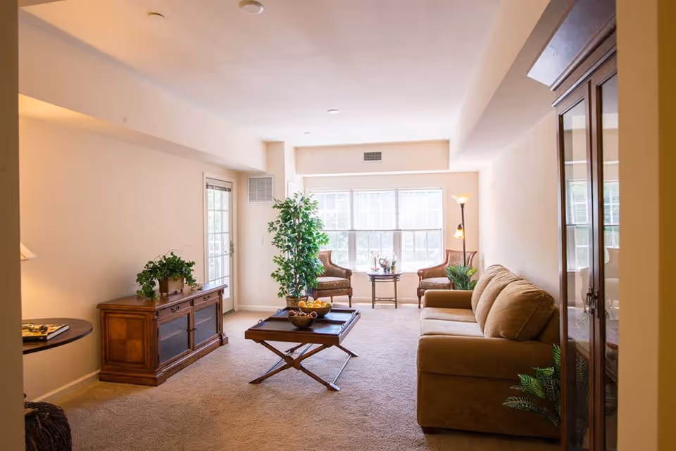 A bright living room with beige walls and carpet, featuring a brown sofa on the right, a wooden coffee table with a bowl of fruit in the center, two armchairs near a large window with blinds, a tall floor lamp, and wooden furniture including a cabinet and a side table with green plants.