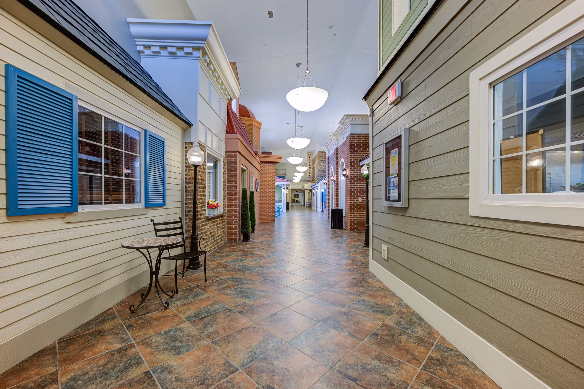 Indoor hallway designed to resemble an outdoor street with building facades on either side, featuring windows, shutters, a small table with a chair, decorative street lamps, and hanging ceiling lights.