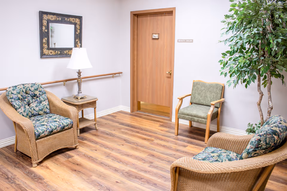 A bright waiting area with wicker chairs, a side table and lamp, a wooden door labeled "Beauty Salon," and a potted tree.