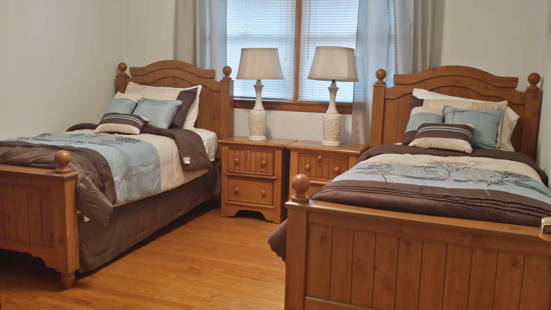 A bedroom with two wooden twin beds, each made with blue and brown bedding and multiple pillows. Between the beds are two wooden nightstands with white ceramic lamps. A window with blinds and light-colored curtains is behind the nightstands.