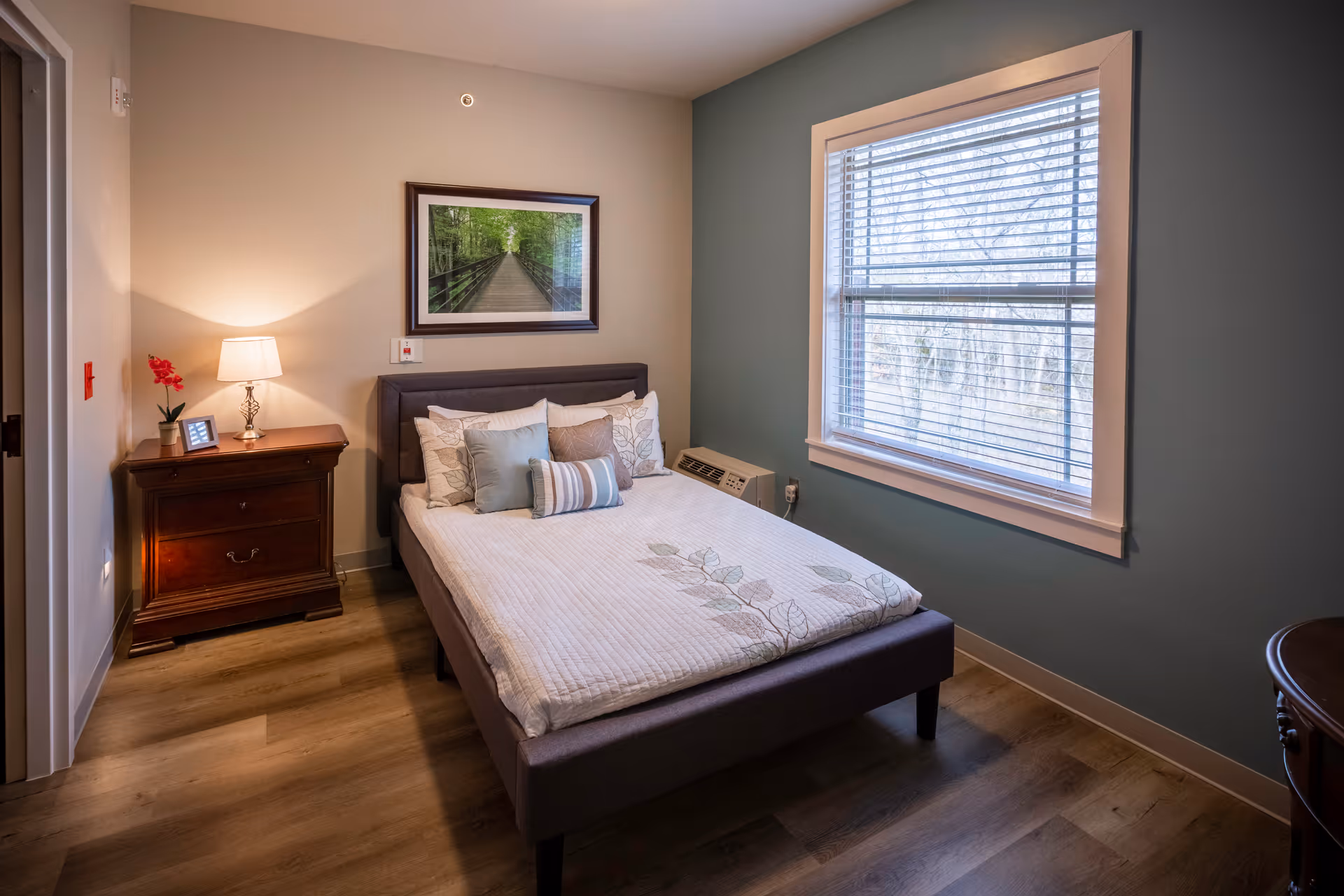 A cozy bedroom with a neatly made bed featuring multiple pillows and a floral-patterned quilt. There is a wooden nightstand with a lamp, a small plant, and a framed photo. A large window with blinds lets in natural light, and a framed picture of a forest pathway hangs on the wall above the bed. The room has wood flooring and walls painted in light beige and muted blue tones.