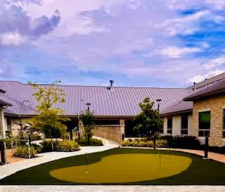 Outdoor courtyard area at Ignite Medical Resort of Katy featuring a putting green surrounded by a paved walkway, small trees, and a building with a metal roof under a partly cloudy sky.