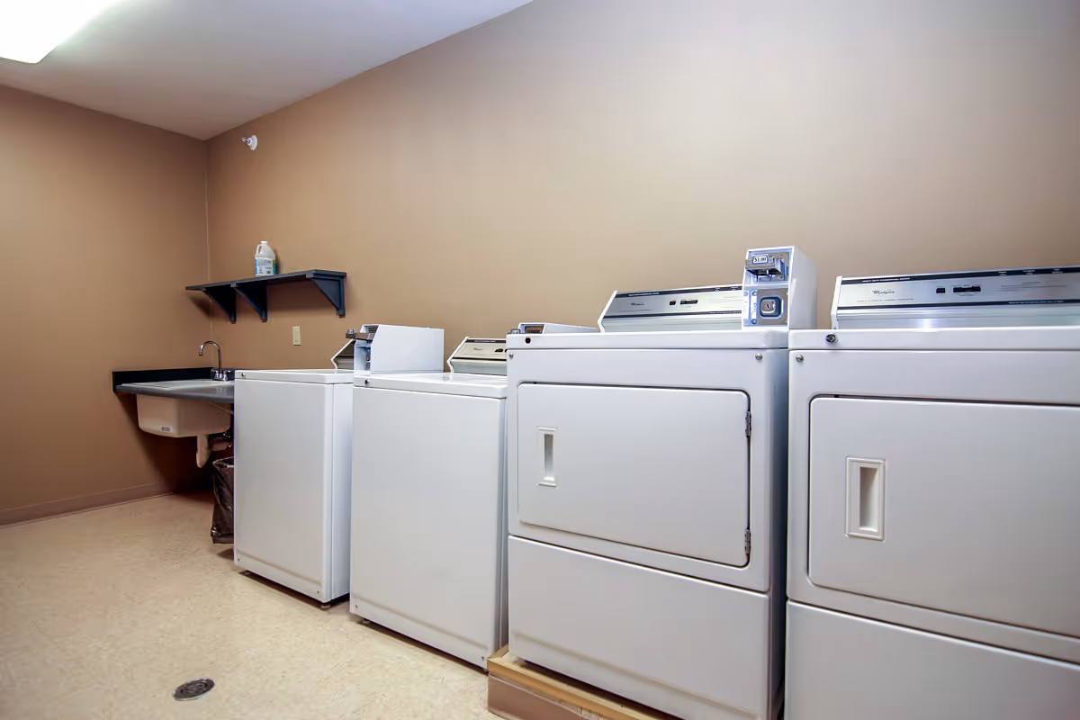 Laundry room with two white washing machines and two white dryers lined up against a beige wall. There is a utility sink with a faucet on the left side and a small black shelf mounted above it holding a bottle of detergent.