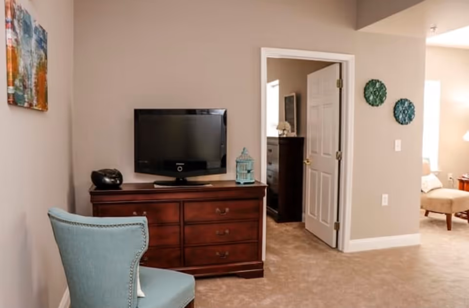 Interior view of a room with beige walls and carpeted floor, featuring a wooden dresser with a flat-screen TV and decorative items on top, a light blue upholstered chair in the foreground, an open door leading to another room with a dresser, and part of a living area with a beige armchair and side table visible in the background.