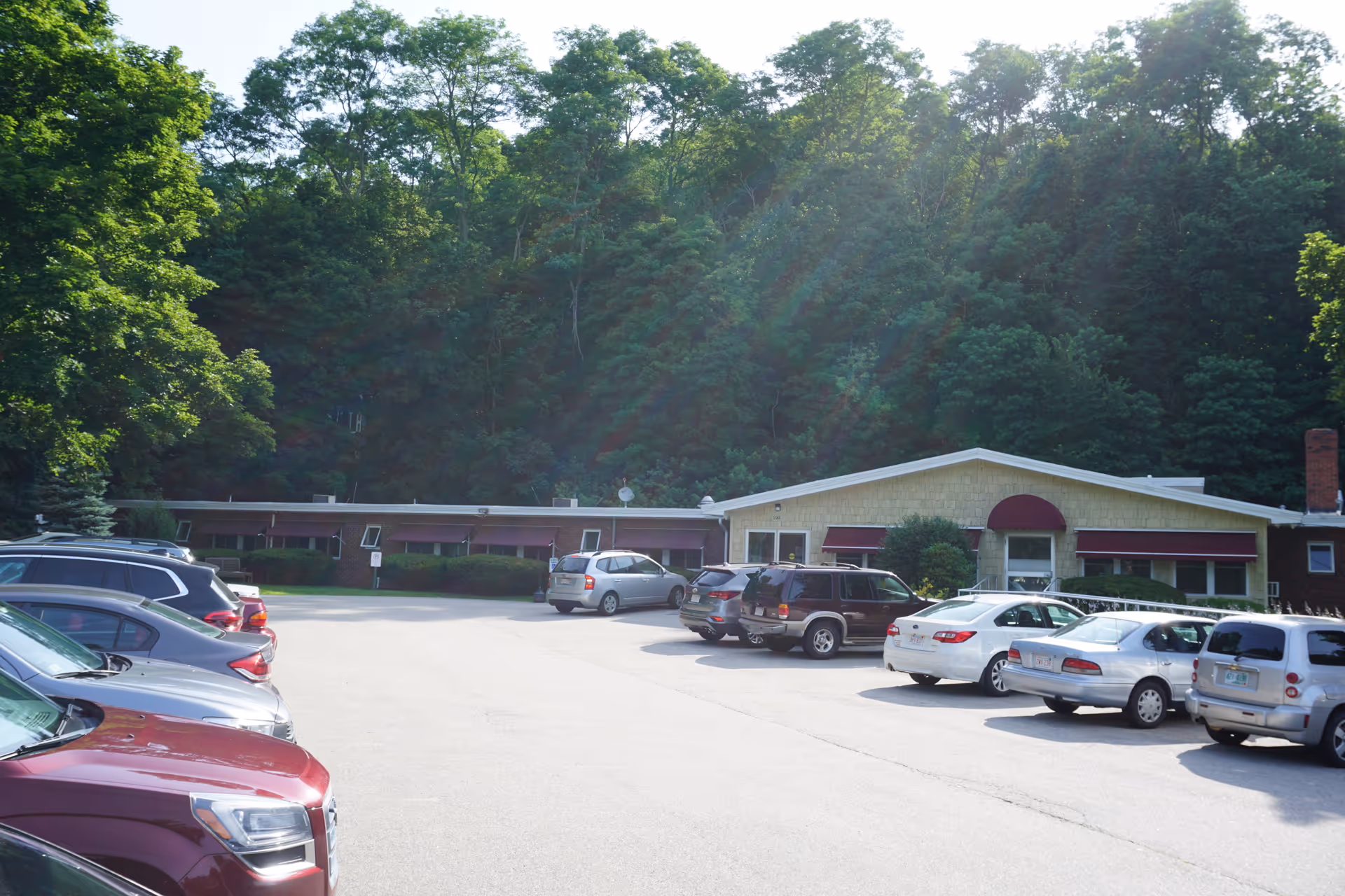 Parking lot with several parked cars in front of a single-story building with maroon awnings, surrounded by dense green trees and forested area in the background.