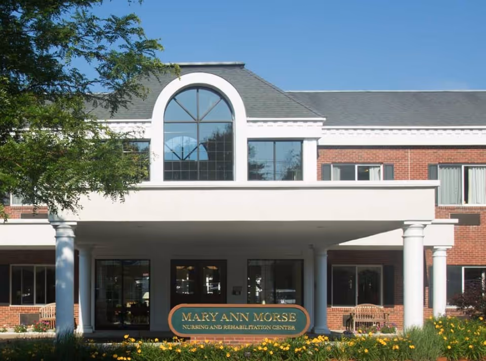 Front exterior view of the Mary Ann Morse Nursing and Rehabilitation Center building with a covered entrance supported by white columns, large windows, and a garden with yellow flowers in the foreground.