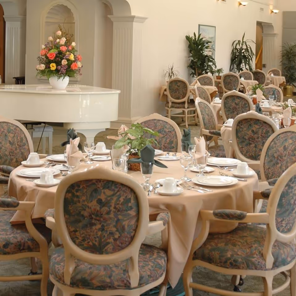 Elegant dining room with round tables covered in beige tablecloths, set with white plates, cups, glasses, and folded napkins. Upholstered chairs with floral patterns surround the tables. A white grand piano with a colorful flower arrangement on top is visible in the background, along with potted plants and classical architectural details on the walls.