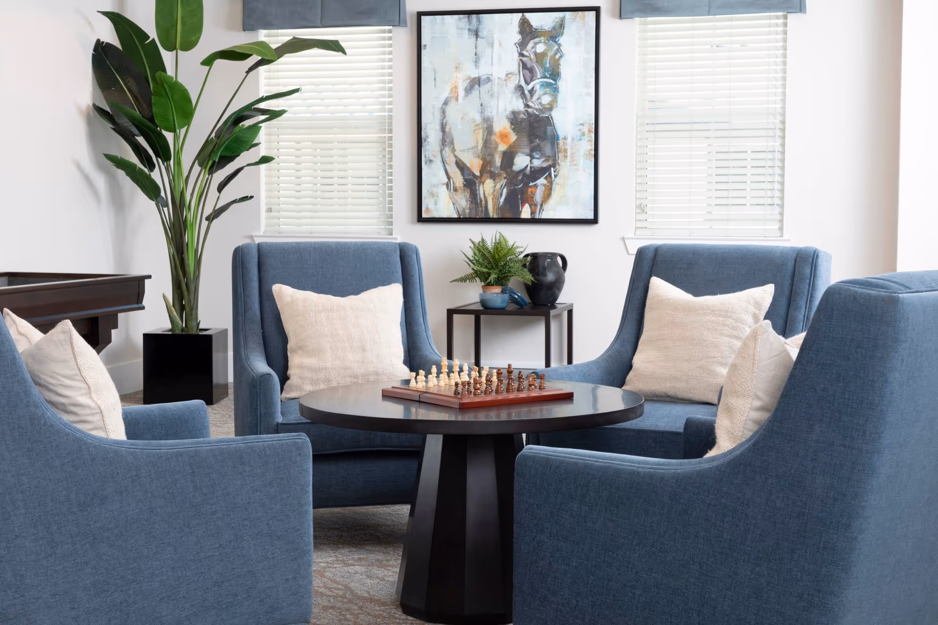 Four blue upholstered armchairs arranged around a round table with a chess set in a bright sitting area decorated with pillows, plants, and framed artwork.