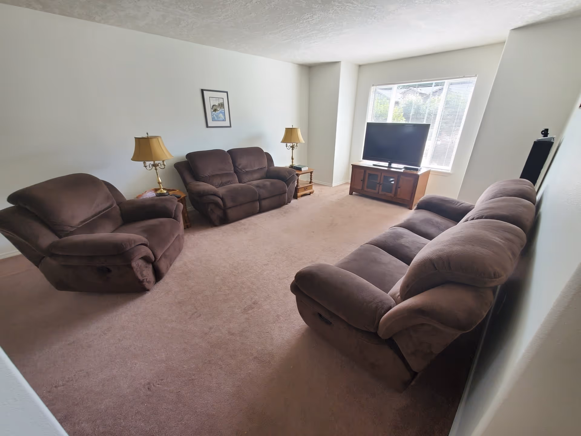 A living room with three brown upholstered recliner sofas arranged around a TV on a wooden stand near a window with blinds. There are two side tables with lamps and a framed picture on the wall.