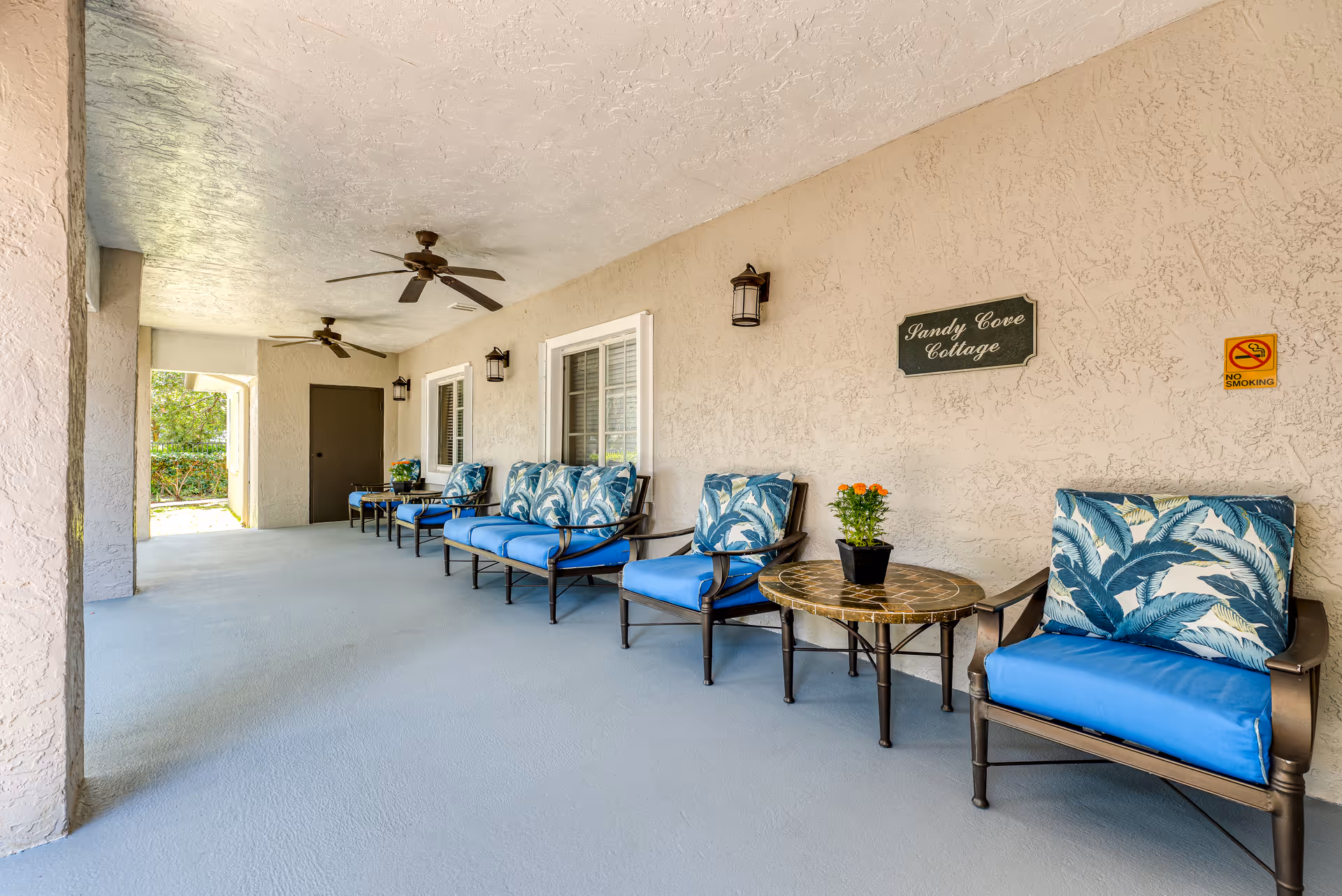 Covered outdoor patio area with several cushioned chairs and small tables arranged along a textured beige wall. Ceiling fans are mounted overhead, and there are wall-mounted lantern-style lights. A sign on the wall reads 'Sandy Cove Cottage' and a no smoking sign is also visible. The patio floor is smooth and painted light blue.